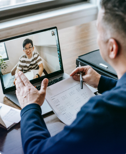 Person in a blue jacket participates in a video conference with a young man in a striped shirt, with notes and a laptop on the desk