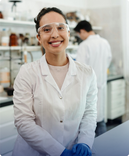 Smiling scientist in a white lab coat and safety glasses, standing in a laboratory with another researcher in the background