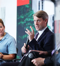 A man in a suit speaks into a microphone during a panel discussion, gesturing with his hand. Two other people listen attentively. The background is blurred.