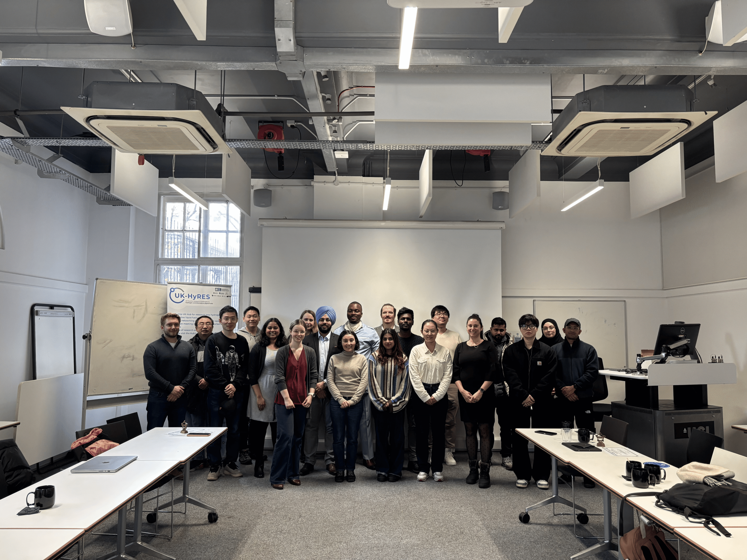 Group of ECR's and PhD students standing infront of U-shaped tables and a whiteboard smiling for a group photo